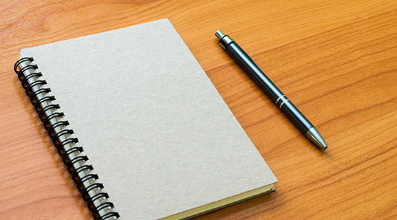 A blank spiral notebook and a pen on a wooden desk.