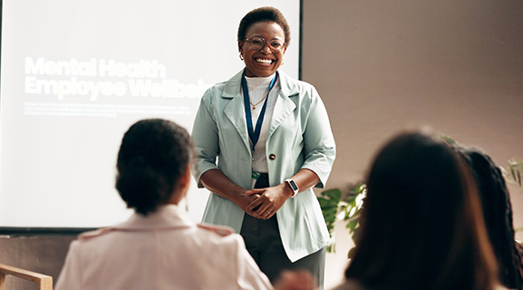 A smiling woman in a light jacket speaks to an audience during a mental health workshop.